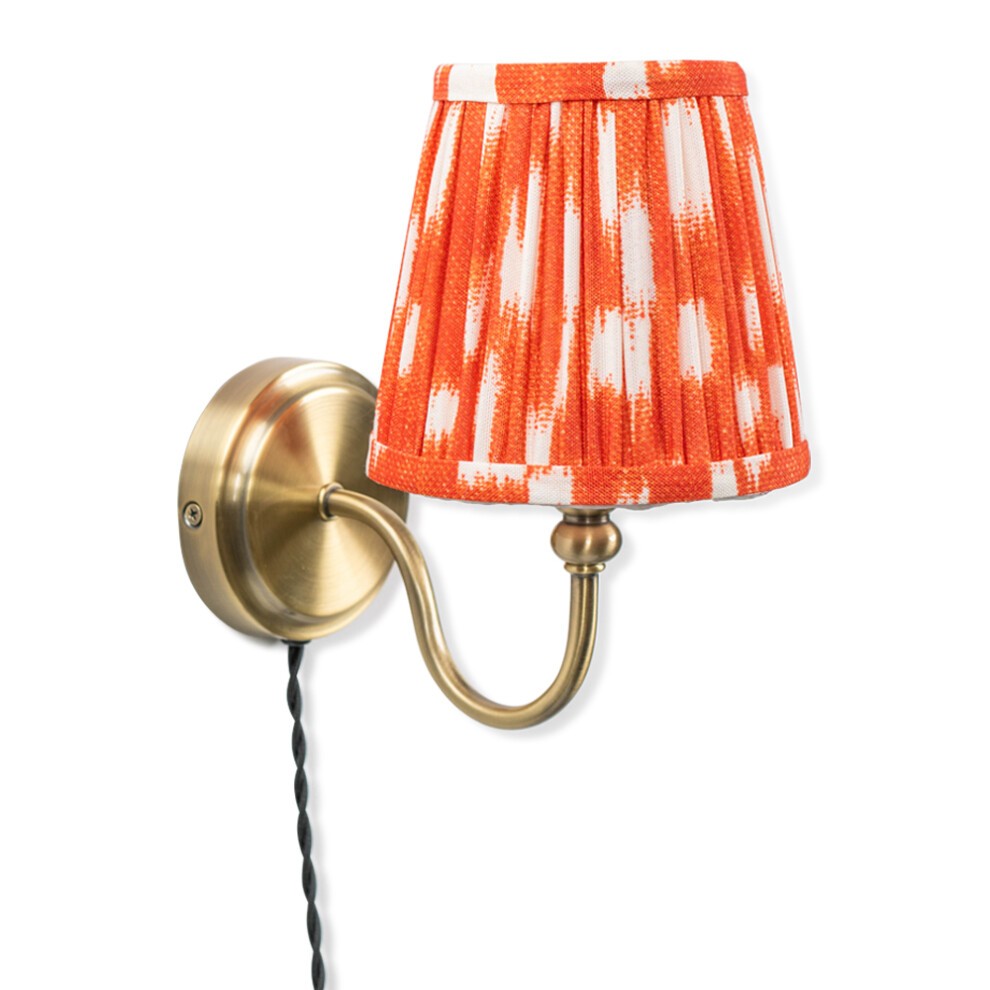 Antique brass wall light featuring an orange pleated tapered shade with white accents, mounted on a curved arm with a black cord.
