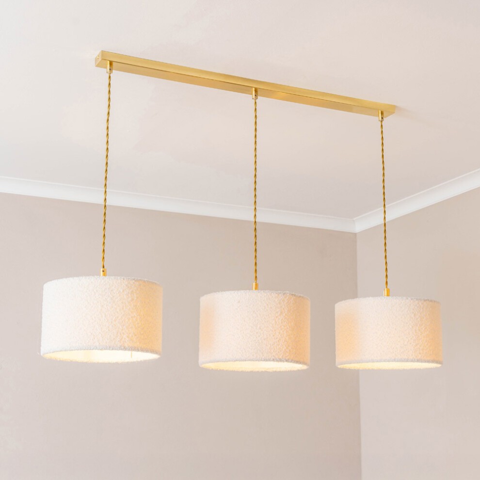Three cream boucle drum shades hang from a gold drop bar, featuring a sleek design with LED bulbs, set against a soft beige background.