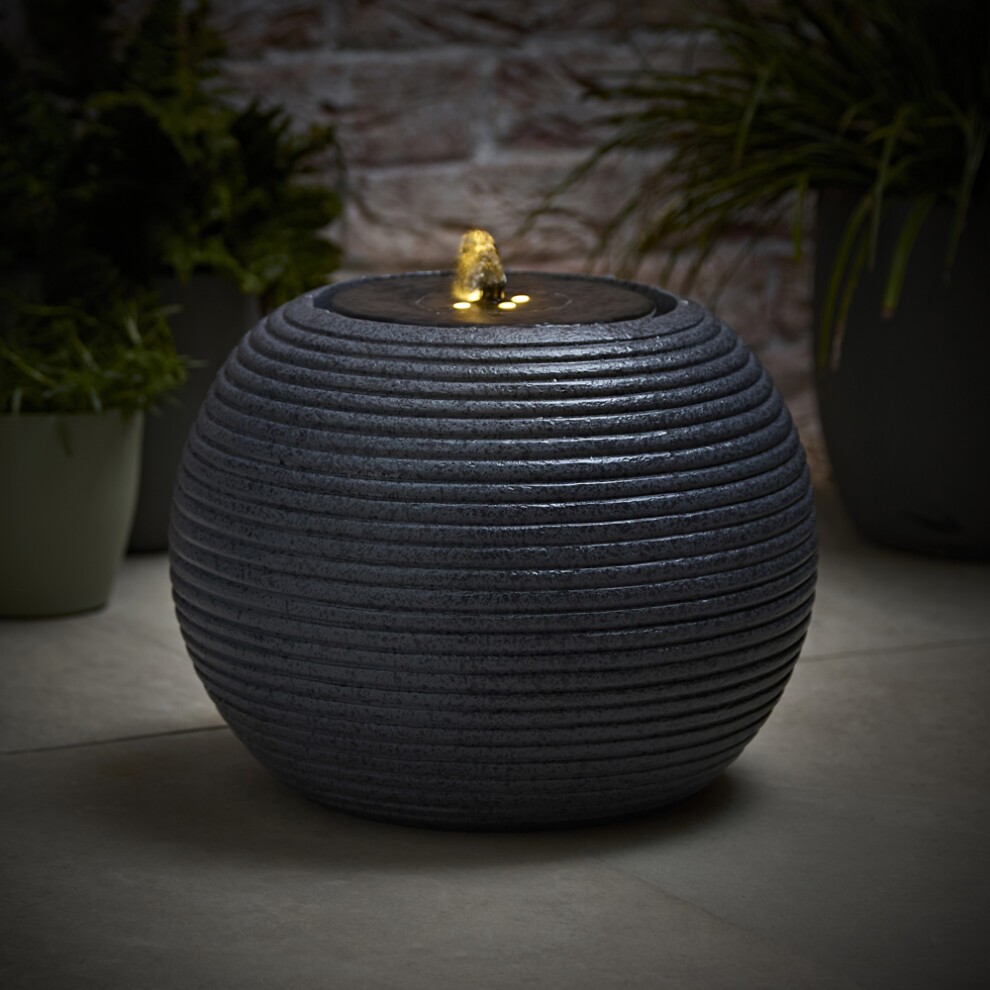 Ribbed grey solar-powered outdoor water fountain with a round design, featuring a gentle water spout and illuminated top, surrounded by potted plants.