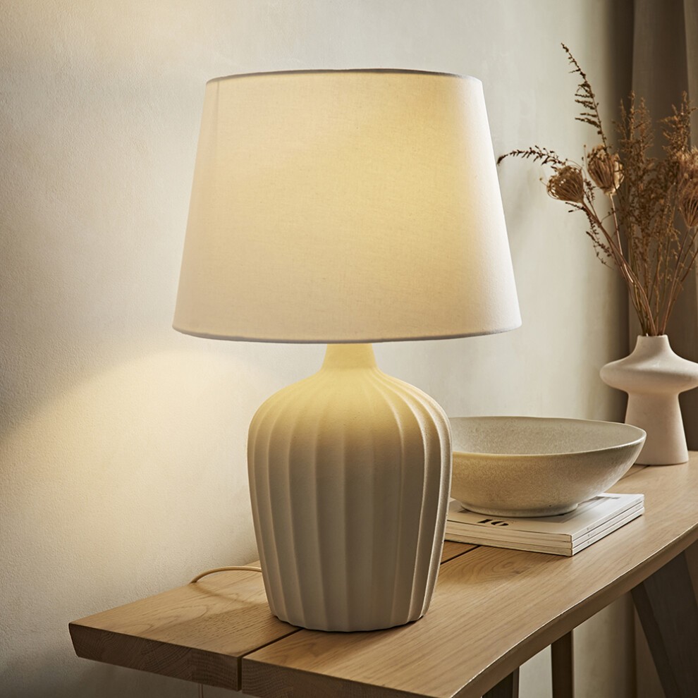 Beige ribbed ceramic table lamp with a natural tapered linen shade, softly illuminated, placed on a wooden table beside decorative items and dried flowers.