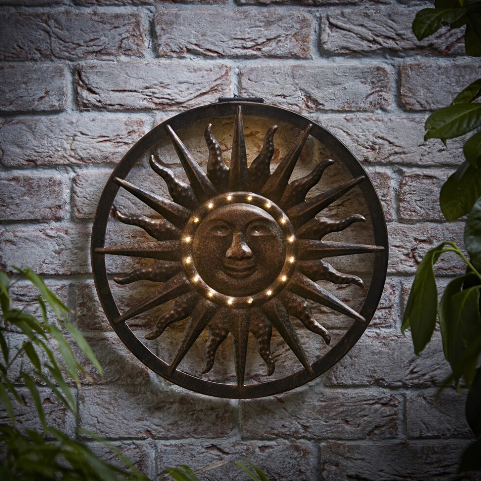 Grey solar-powered wall art featuring a sun design with a smiling face and rays, illuminated by small lights, mounted on a brick wall surrounded by greenery.
