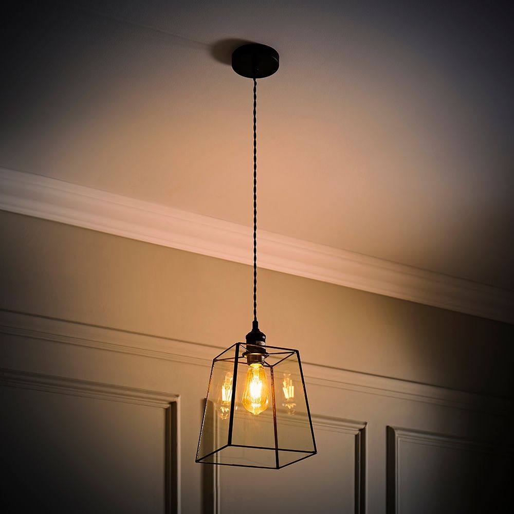 Black metal pendant light with a geometric glass lantern shade, featuring a warm bulb, suspended from a ceiling mount with a braided cord against a neutral wall.