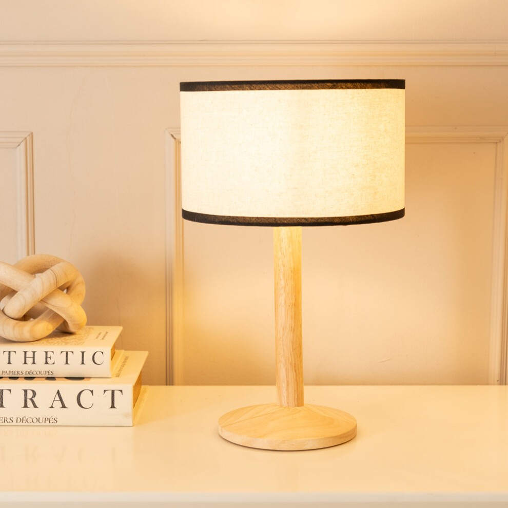 Natural light wood table lamp with a cylindrical linen shade featuring a black trim, resting on a round base, illuminating a softly styled interior.