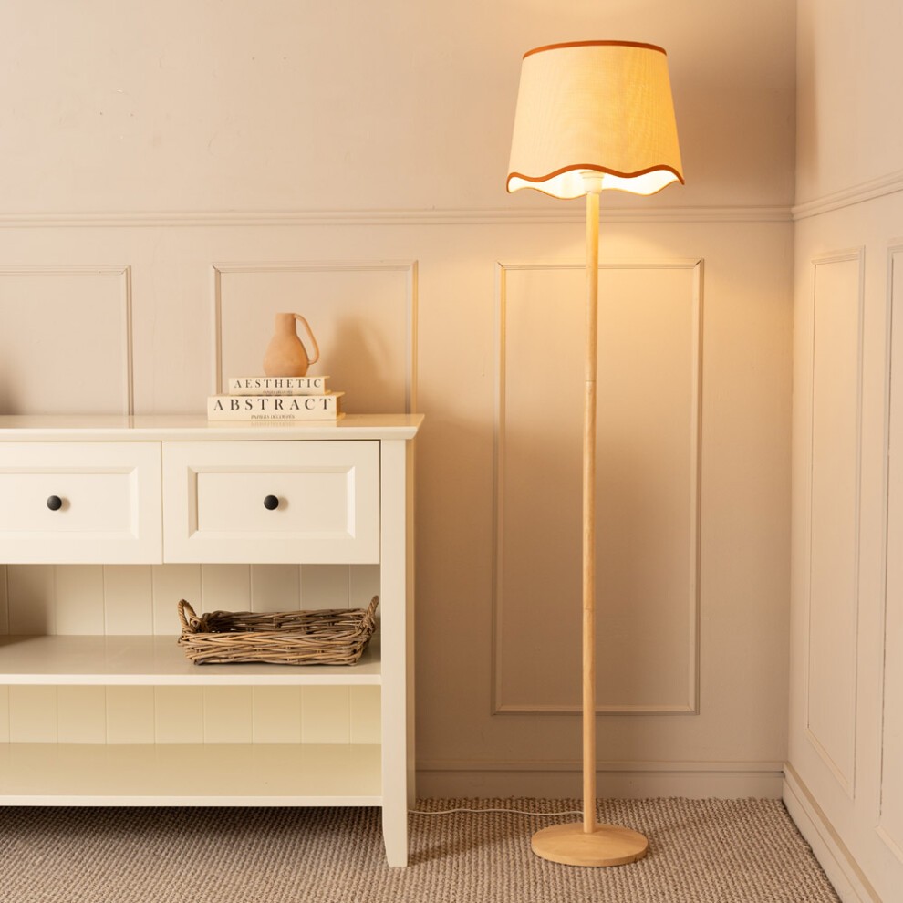 Natural wood floor lamp with a tapered scallop trim shade in burnt orange, featuring an LED bulb, positioned beside a white cabinet and decorative items.