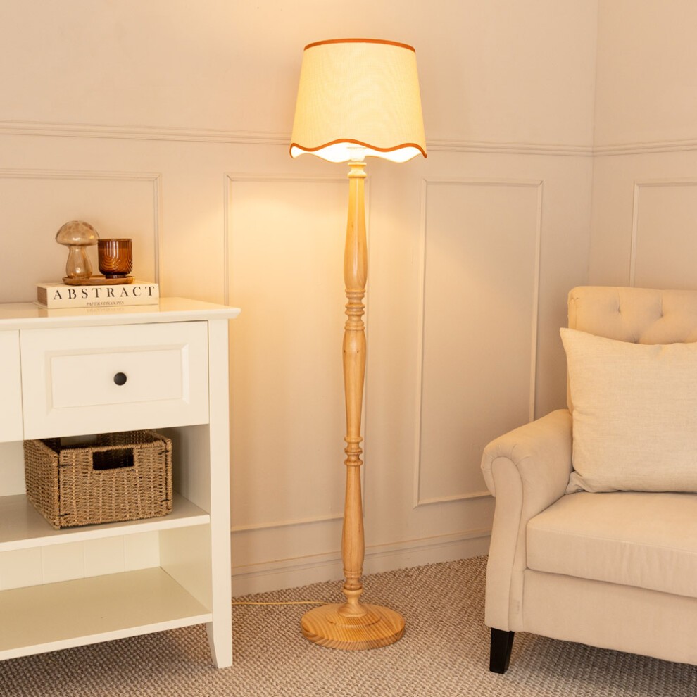 Tall light wood floor lamp with a tapered linen shade featuring a scallop trim in burnt orange, positioned beside a white side table and a beige sofa.