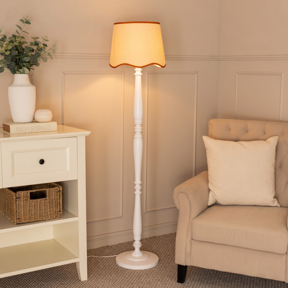 White wood floor lamp with a tapered linen shade featuring scallop trim in orange, standing beside a beige armchair and a white side table with a plant and decorative items.