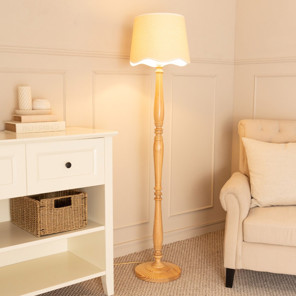 Tall light wood floor lamp with a tapered linen shade featuring scallop trim, positioned beside a white shelf and beige upholstered chair in a cozy room setting.