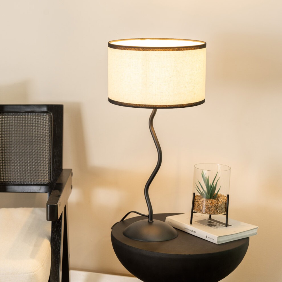 Wiggle black metal stem table lamp featuring a beige linen shade with black trim, placed on a round black table beside a glass planter with a small plant.