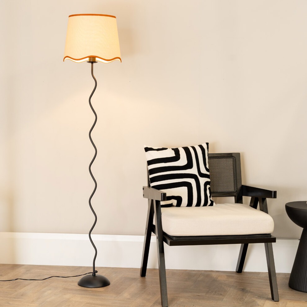 Wiggle black metal floor lamp with a tapered linen shade featuring burnt orange scallop trim, standing beside a black and white striped cushion on a modern chair.