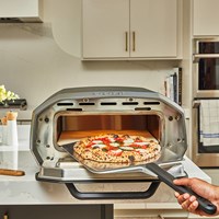 Silver Ooni Volt 12 Electric Oven with a wooden pizza peel, showcasing a freshly baked pizza topped with tomatoes and cheese, set on a modern kitchen countertop.