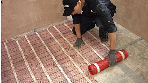 A person in black clothing lays down a red and white underfloor heating mat on a concrete floor, showcasing the mat's grid pattern and adhesive backing.