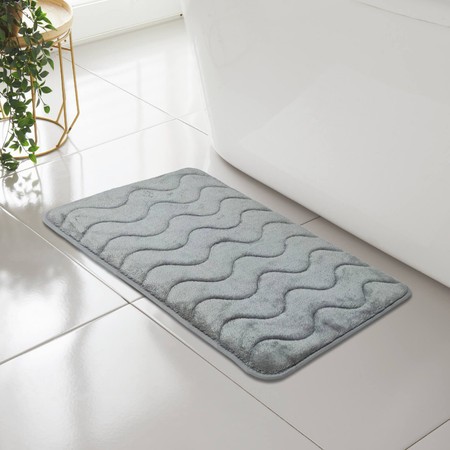 Soft, wave-patterned memory foam bath mat in a calming light gray color, featuring a plush texture and a non-slip backing, placed next to a white bathtub on a tiled floor.