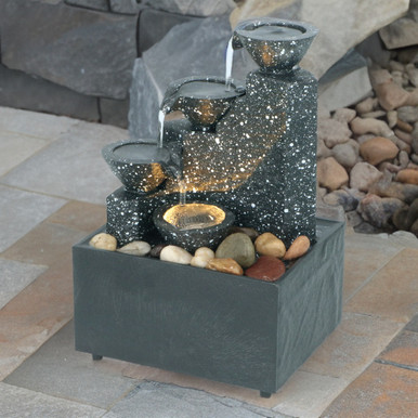 Grey three-tier water fountain featuring a textured stone finish, cascading water from bowls, illuminated base, and surrounded by smooth pebbles.