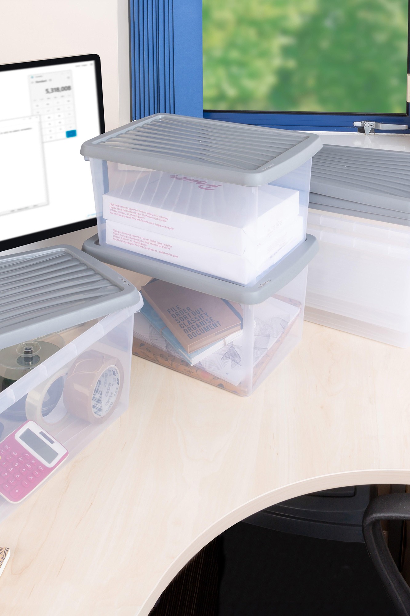 Stackable transparent plastic storage boxes with gray lids, featuring a ribbed design, filled with office supplies, placed on a light wooden desk near a computer.