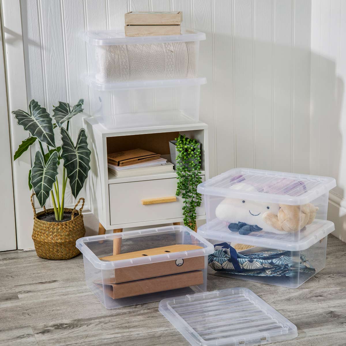 Clear plastic storage boxes with lids, featuring a stackable design, placed beside a white cabinet and a potted plant, showcasing various items inside, including textiles and toys.