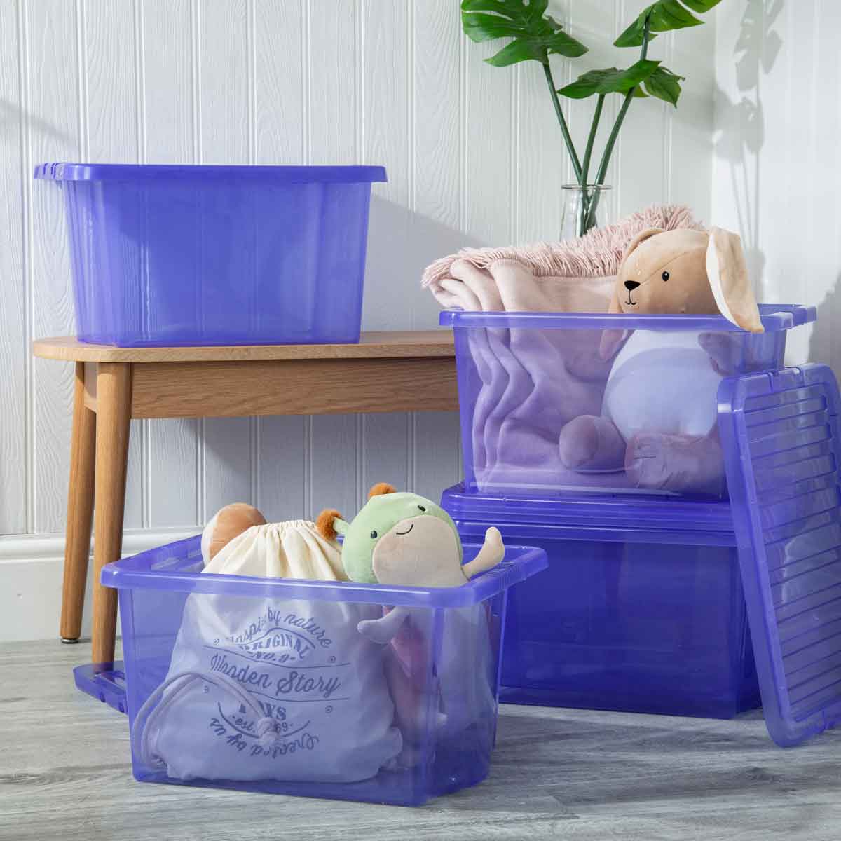 Transparent blue plastic storage boxes with lids, featuring a textured design, stacked and filled with toys and blankets, placed beside a wooden table and a potted plant.
