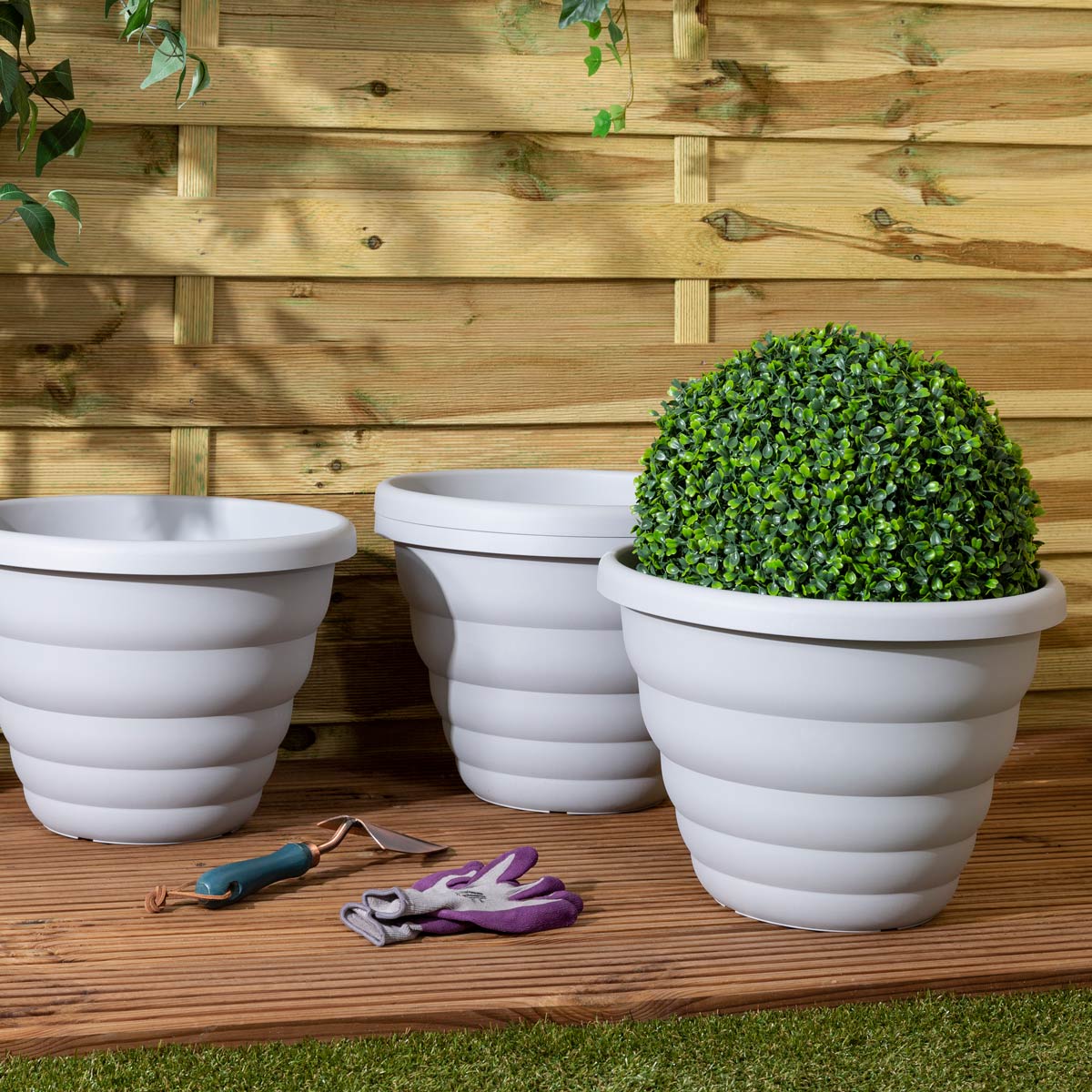 Set of three 40cm round beehive pots in cement grey, featuring a ribbed design, with a faux topiary in the largest pot, placed on a wooden surface against a fence.