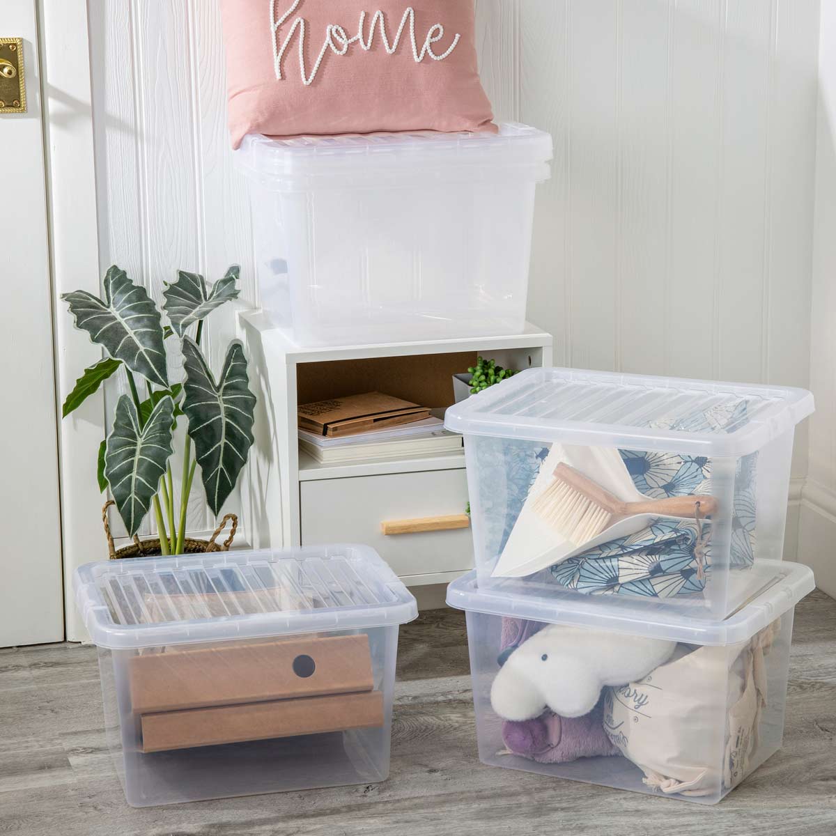 Clear plastic storage boxes with lids, stacked and filled with various items, including soft toys and storage boxes, set against a light-colored wall and greenery.