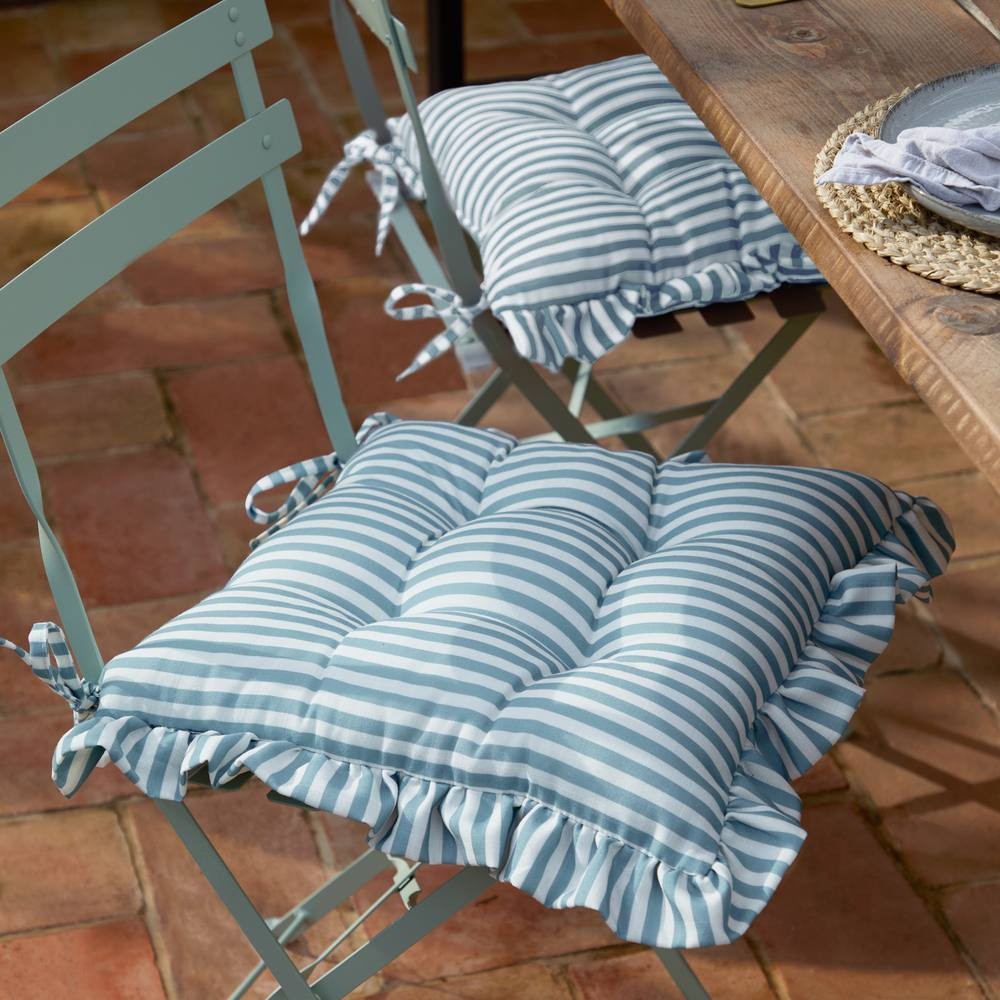 Blue and white striped ruffle seat pads with ties, designed for indoor or outdoor use, placed on light blue metal chairs atop a terracotta tiled surface.