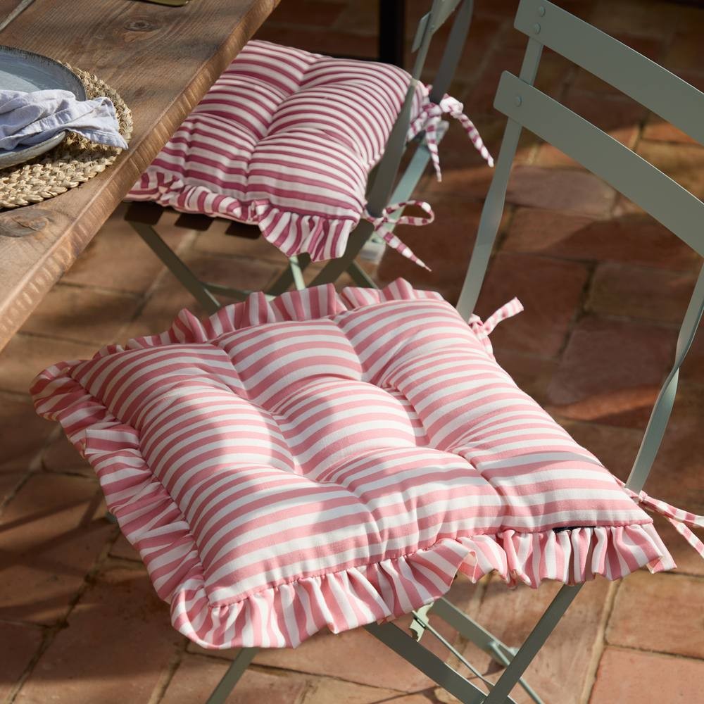 Pink and white striped seat pads with ruffles, featuring ties for attachment, designed for indoor or outdoor use, placed on a rustic wooden table with metal chairs.