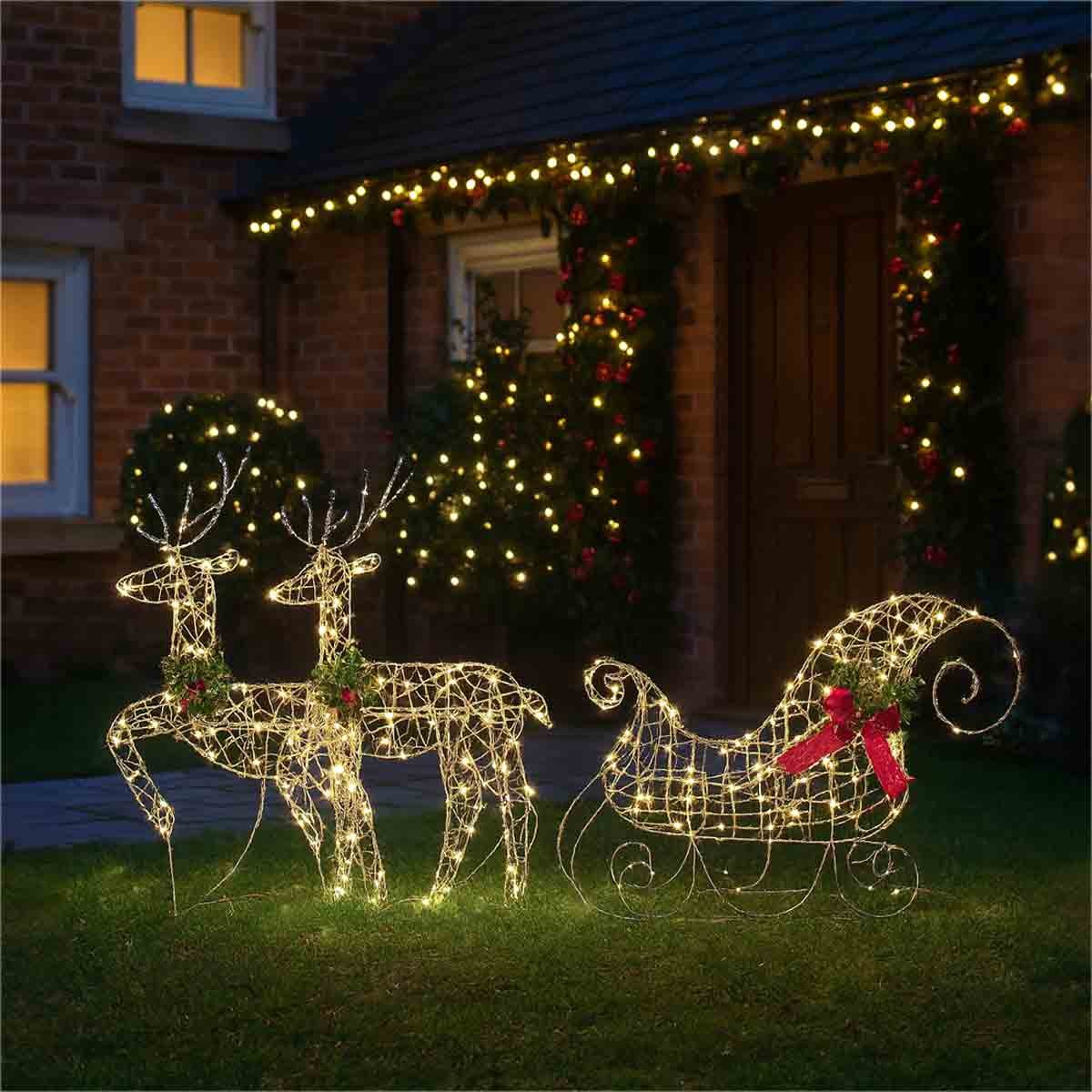 Two silver LED deer with antlers and a decorated sledge, all made of wire, illuminated with lights, set on a grassy area in front of a house adorned with festive lights.