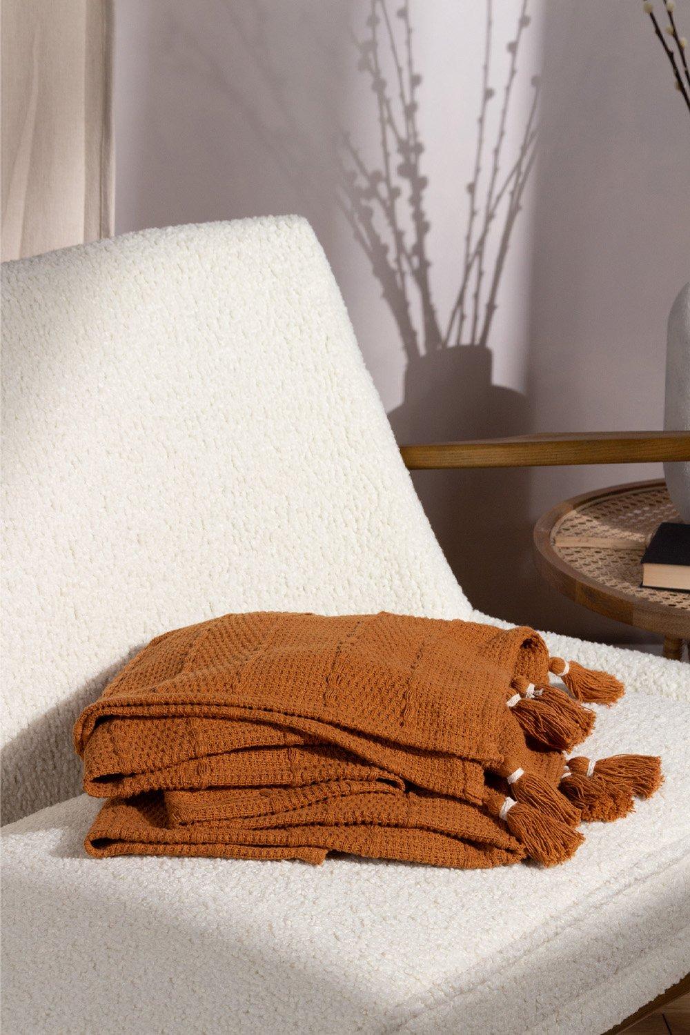 Ginger-colored cotton throw with woven texture and tassels, neatly folded on a light-colored chair, complemented by soft shadows and a decorative vase in the background.