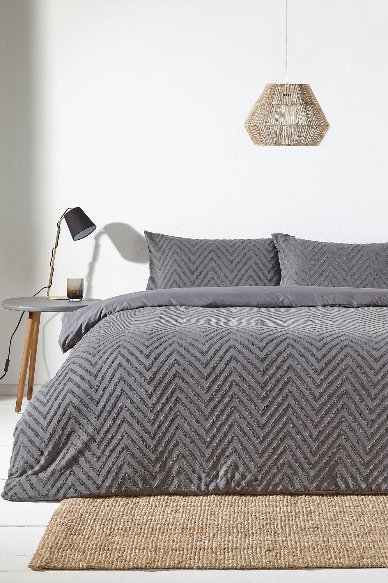 Gray chevron-patterned bedding set with matching pillowcases, accompanied by a woven pendant light, a black lamp, and a round side table on a light wooden floor.