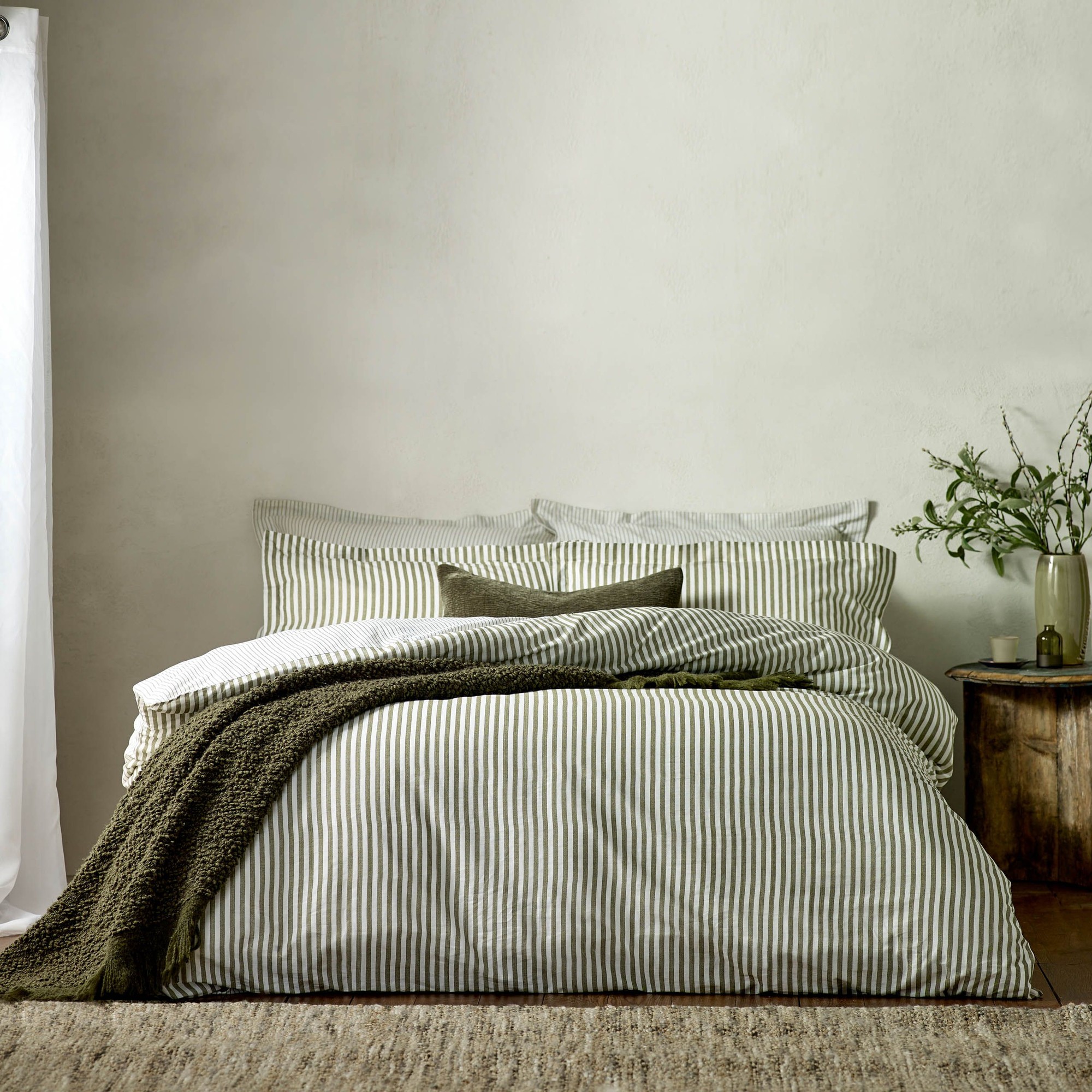 Hebden Mélange Stripe duvet cover set in soft cotton features alternating green and white stripes, complemented by a textured olive throw and decorative pillows on a cozy bed.