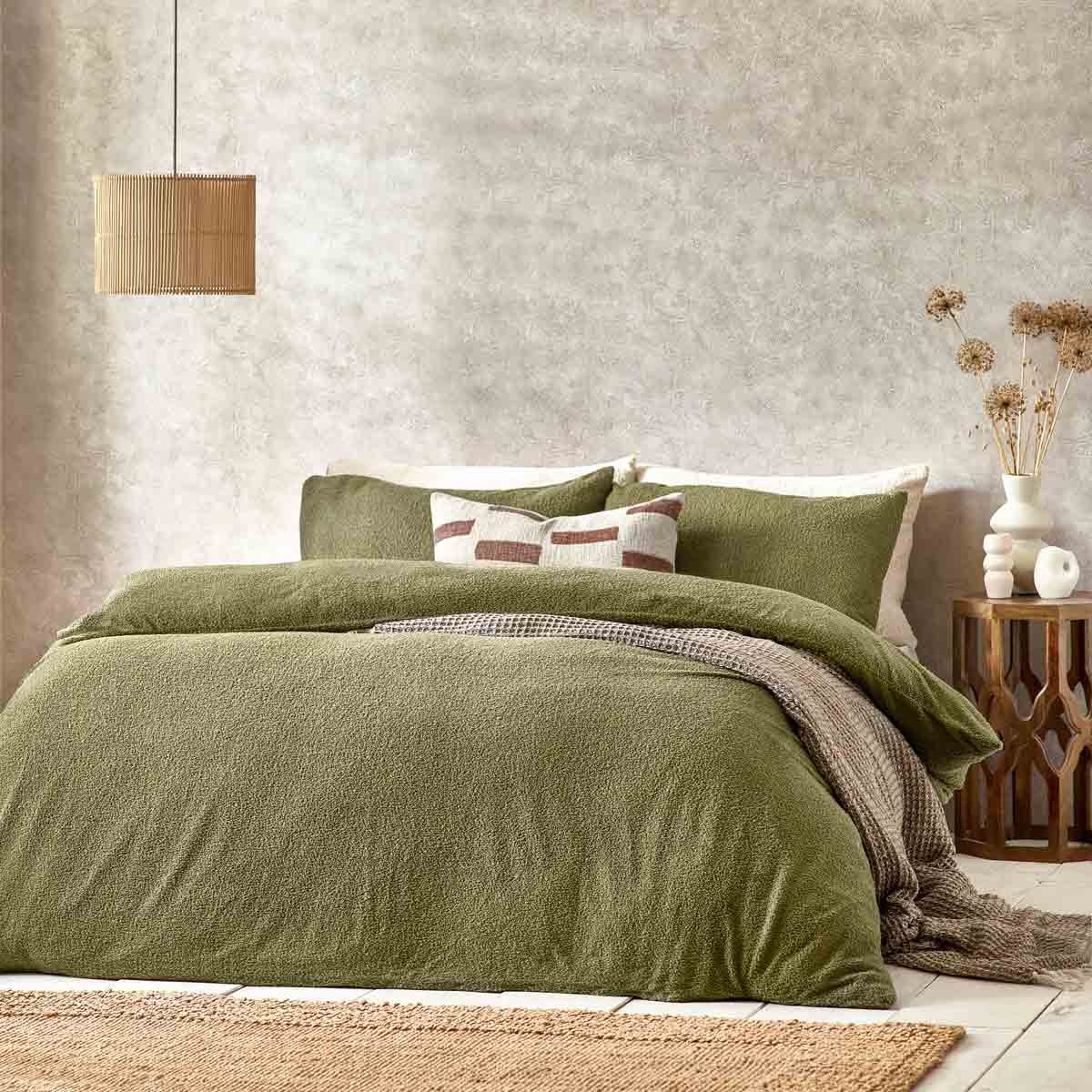 King-sized duvet cover set in olive green boucle texture, featuring matching pillow shams and layered with a patterned throw, set against a neutral, textured wall and natural decor.
