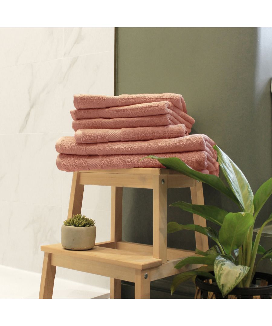 Soft blush cotton towels stacked neatly on a wooden stool, with a small potted plant beside them, against a backdrop of light marble and green wall.