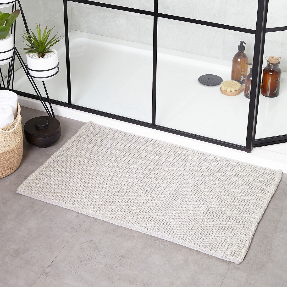 Grey woven cotton bath mat with a loop texture, featuring a non-slip backing, placed on a tiled floor near a shower, surrounded by plants and bath products.