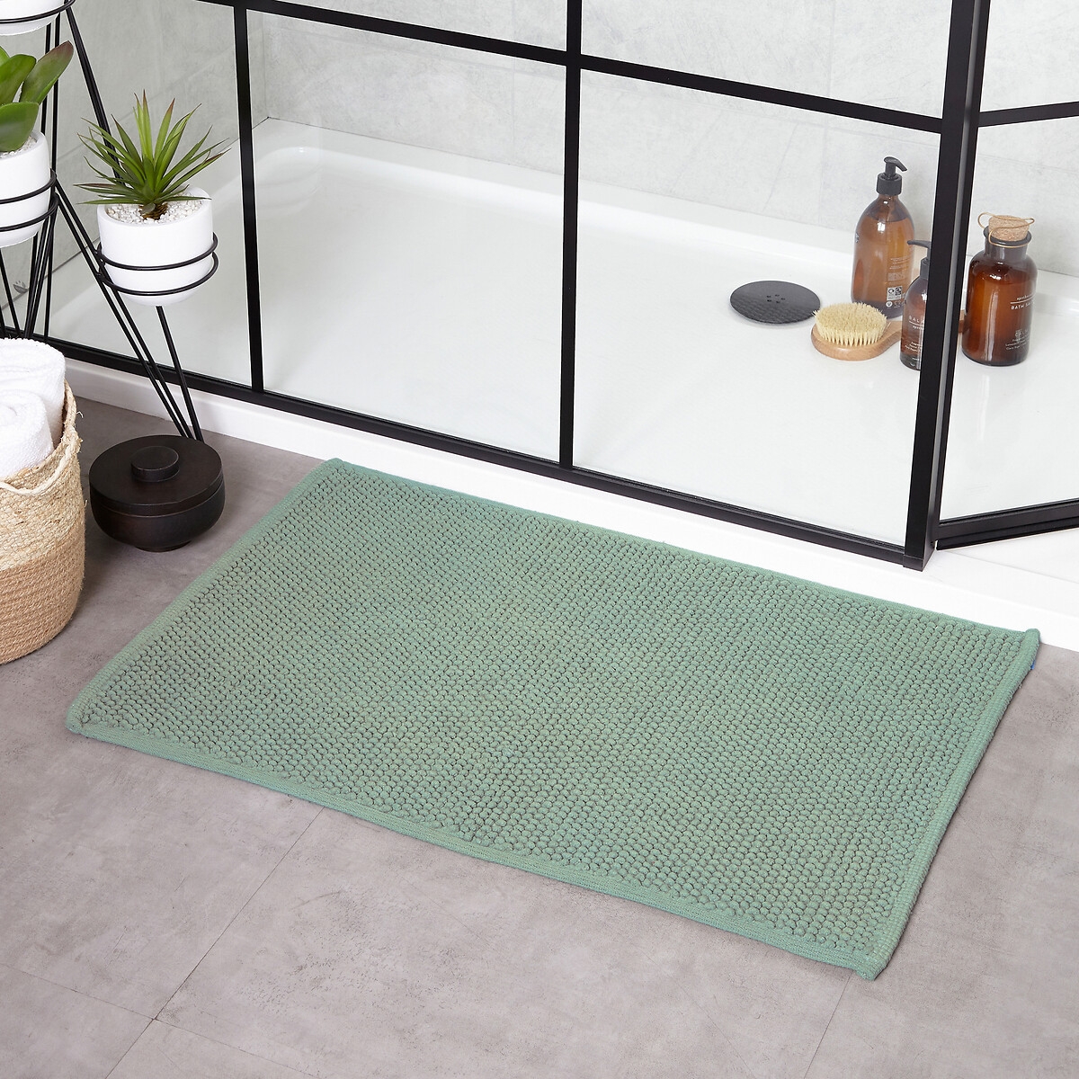 Green woven cotton bath mat with a textured loop design, featuring a non-slip backing, placed on a tiled floor near a glass shower enclosure.