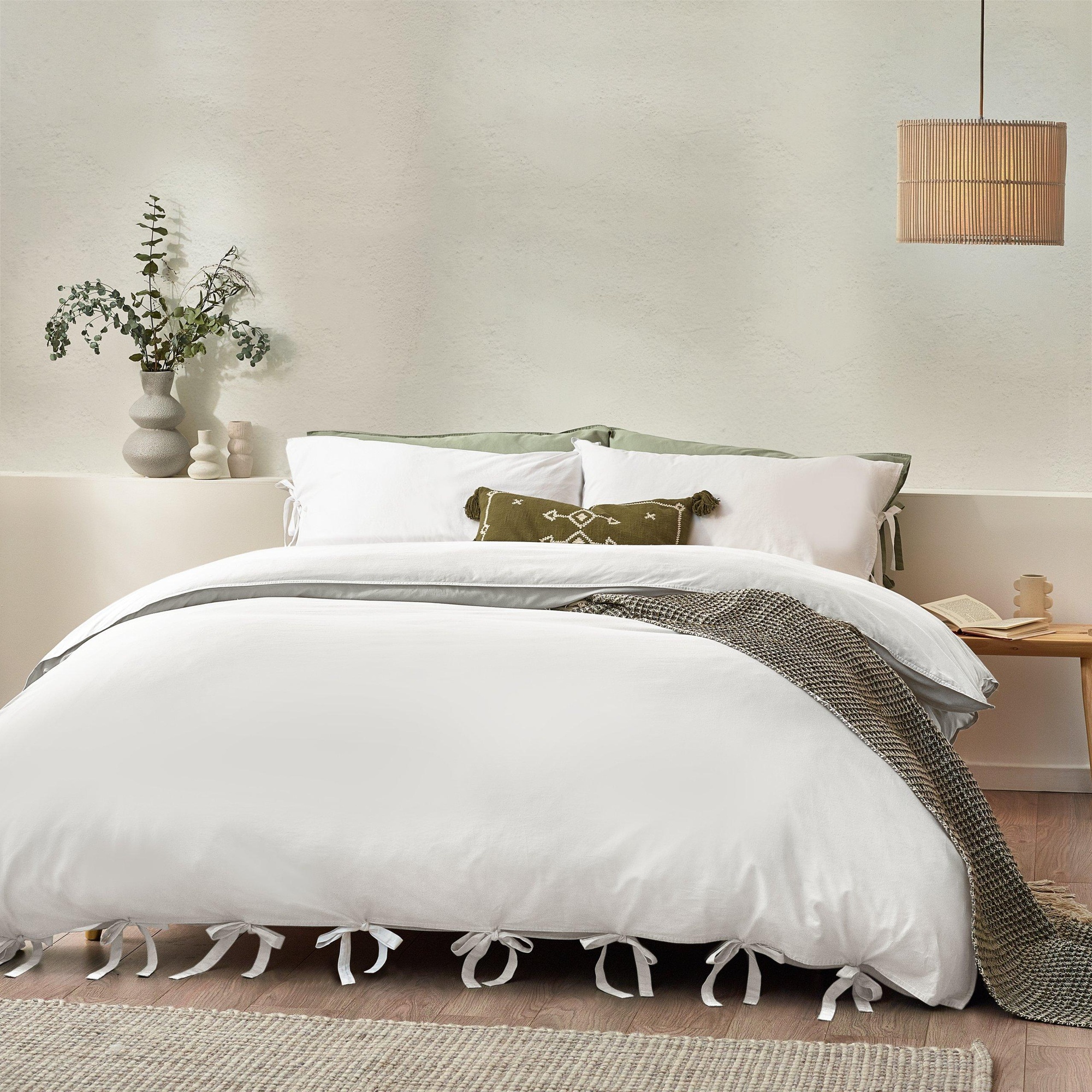 White cotton duvet set featuring a bowtie design, complemented by a green accent pillow, a textured throw, and a minimalist decor with a plant and lamp.