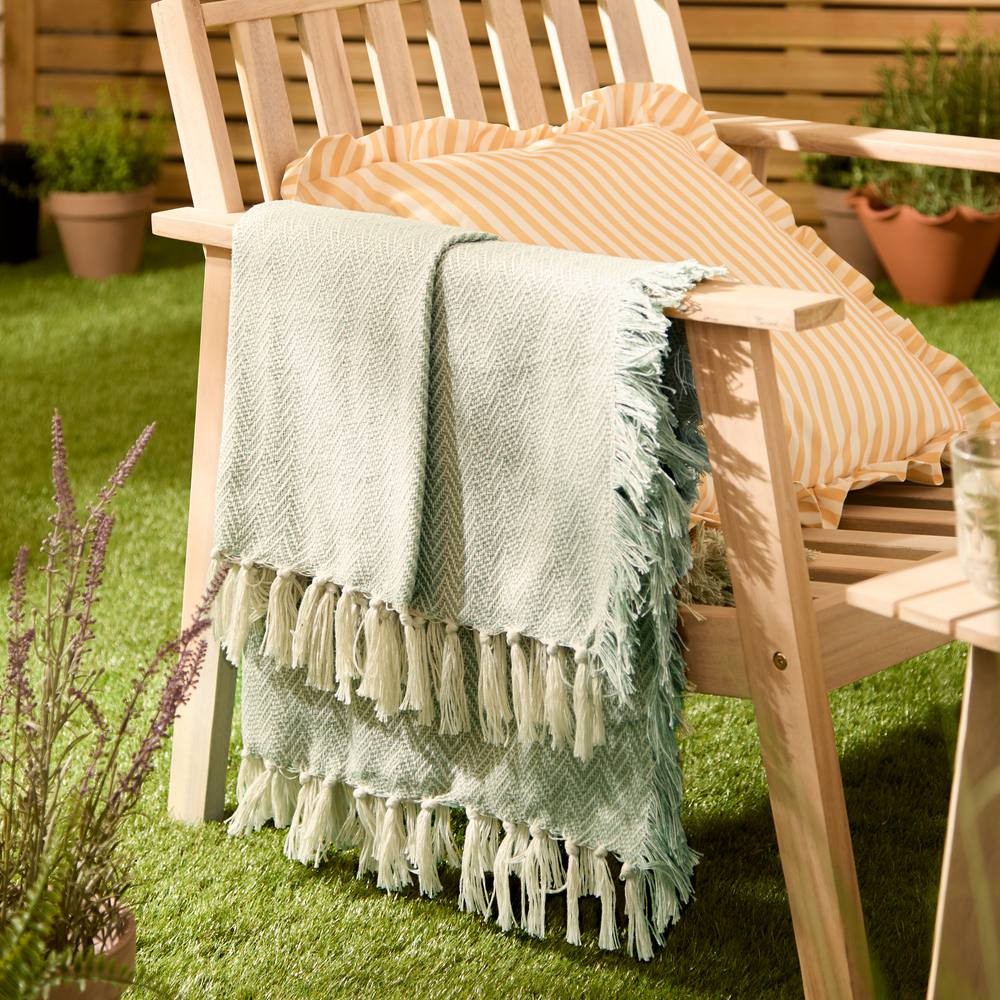 Green woven outdoor throw with fringed edges, draped over a wooden chair, alongside a striped yellow pillow, set on a grassy surface with potted plants in the background.