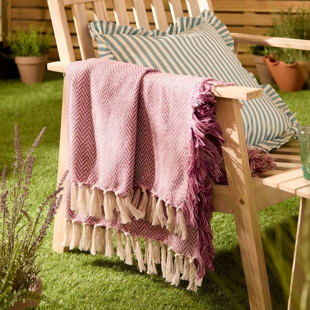 Pink woven outdoor throw with a herringbone pattern and fringed edges, draped over a wooden bench beside a striped cushion, set on green grass.