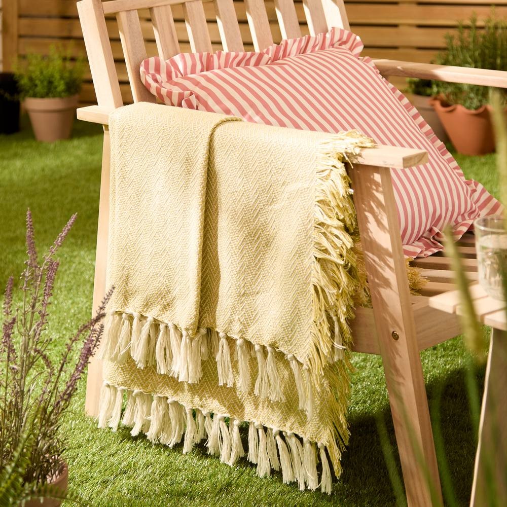 Yellow woven outdoor throw with fringed edges draped over a wooden chair, accompanied by a pink and white striped pillow, set against a grassy background.