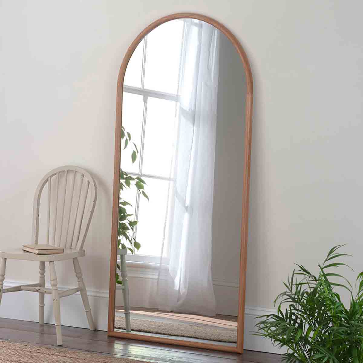 Full-length arched wall mirror featuring a natural oak frame, reflecting light and surroundings, set against a light wall with a chair and indoor plant nearby.