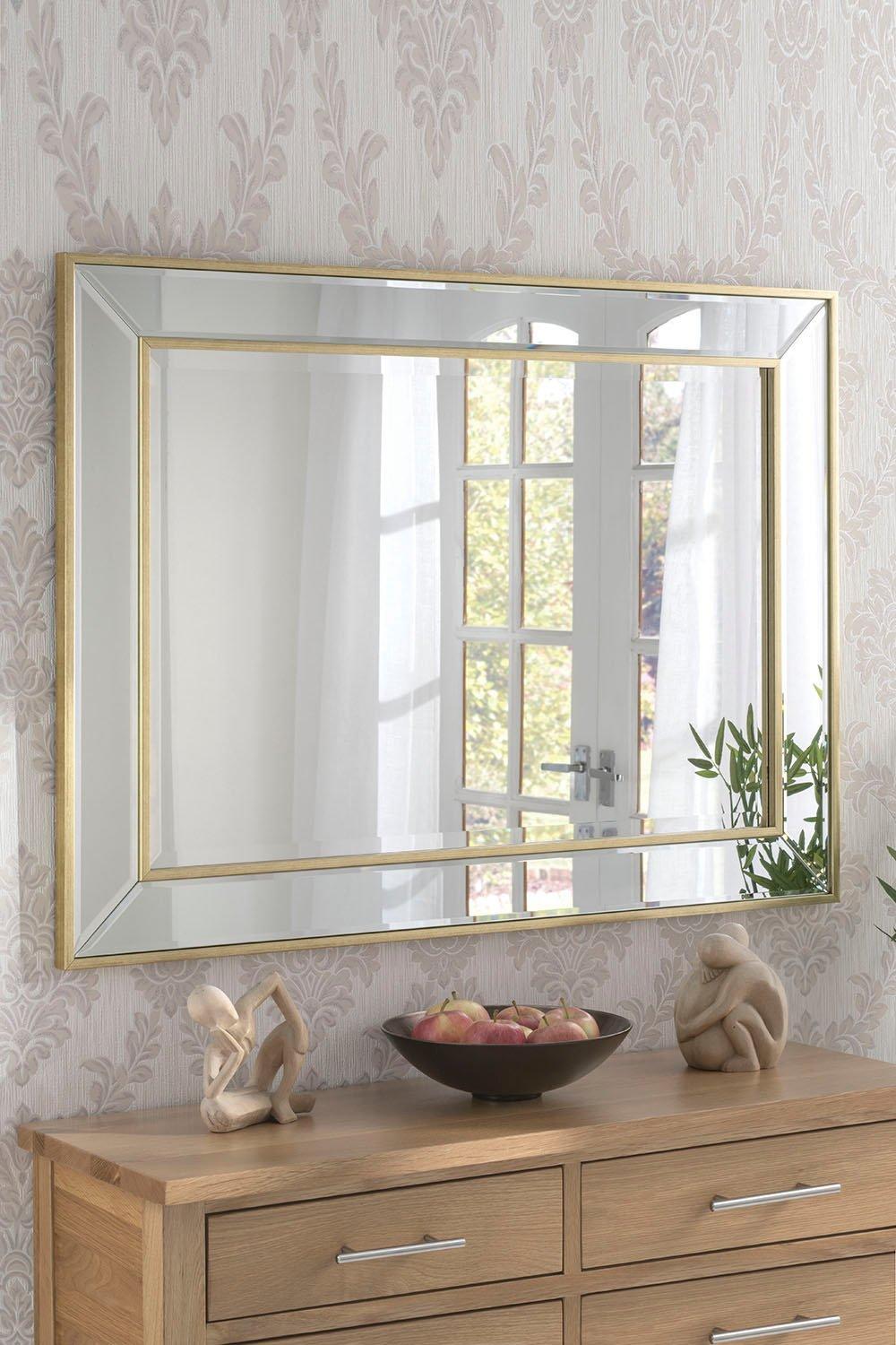 Angled wall mirror in metallic gold with a brass frame, measuring 95x120.5cm, reflecting light and enhancing the room's elegance against a patterned wallpaper backdrop.
