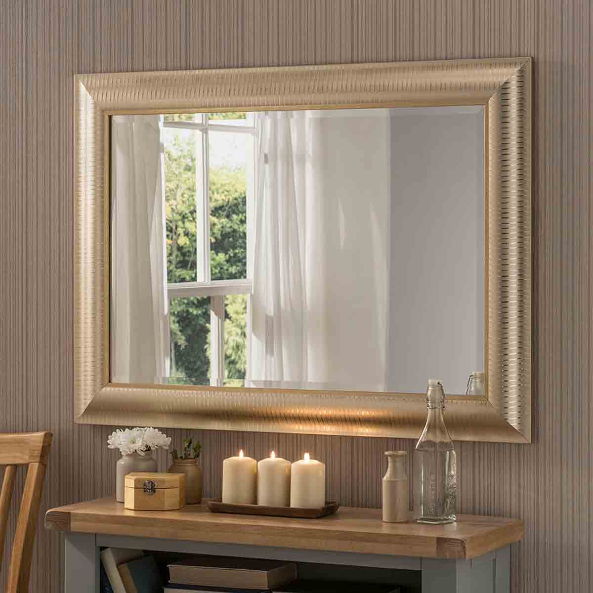 Textured gold bevelled wall mirror with a decorative frame, reflecting natural light, placed above a wooden table with candles and a vase of flowers.