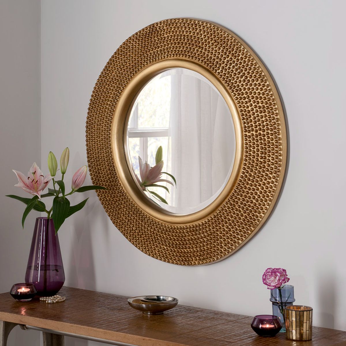 Round bobble mirror in metallic gold, 79cm in diameter, featuring a textured, bubble-like frame, elegantly displayed on a wall above a wooden table with flowers and candles.