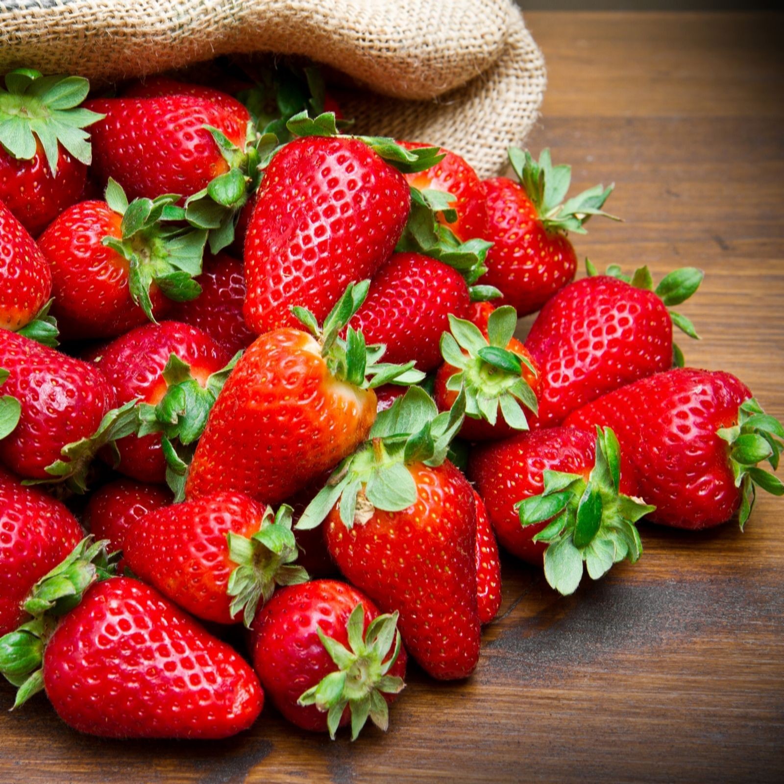 Fresh, vibrant red strawberries with green leafy tops, scattered on a wooden surface, with a burlap sack in the background.