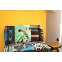 Colorful patchwork sofa with bird and peacock designs against a bright yellow wall, next to a small round gold table.