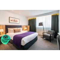 Hotel room with a double bed, purple throw, two bedside lamps, armchair, and large window with dark curtains.