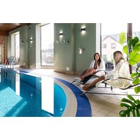 Indoor spa pool with blue water, two women in robes seated on loungers, large windows, and green plants nearby.