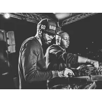 DJ wearing a cap and glasses, performing on stage with another person, surrounded by equipment and stage lights.