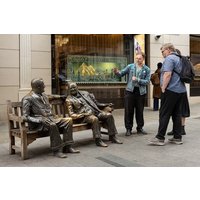 Bronze statues of two seated figures on a bench, with two people standing nearby on a city street.