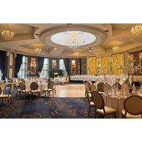 Elegant hotel ballroom with round tables, white tablecloths, gold chairs, chandeliers, and large windows with blue curtains.