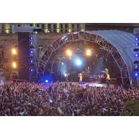 Large outdoor concert stage with arched metal frame, bright spotlights, and a crowd of people in front.