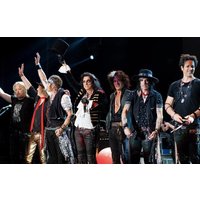 Band on stage with guitars and microphones, wearing rock attire, hats, and scarves, under bright stage lights.