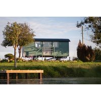 Green tiny house on stilts with large glass windows, black roof, surrounded by trees and grass by a wooden dock.